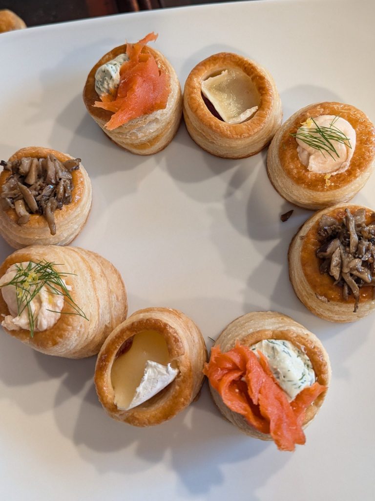 A circular arrangement of pastry cups filled with various toppings, including smoked salmon, cream cheese, and mushrooms, placed on a white plate.